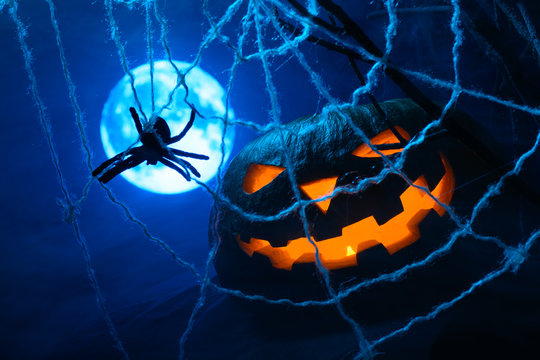 Scary Halloween pumpkin with a glowing face and a cobweb with a spider on the background of the full moon in the fog