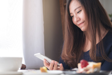 Closeup image of a smiley beautiful Asian woman holding , using and looking at smart phone with white coffee cup and dessert plate on wooden table in vintage cafe