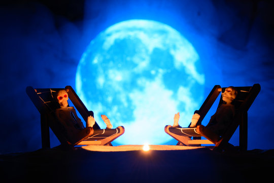 Two Toy Skeleton On The Lounge Chairs Opposite A Burning Candle On The Background Of The Full Moon