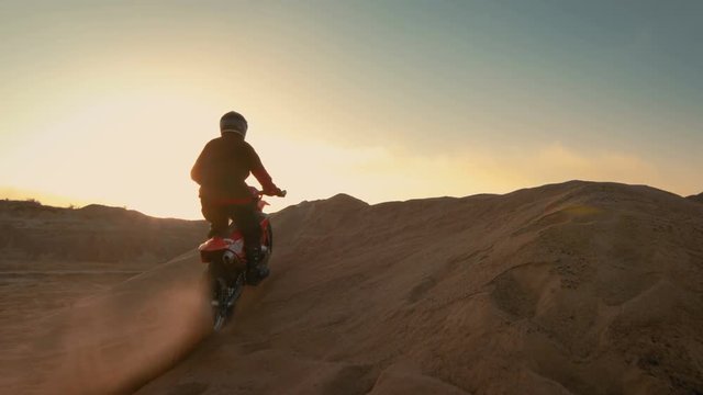 Professional Motocross Motorcycle Rider Driving Over the Dune and Further Down the Off-Road Track. It's Sunset and Track is Covered with Smoke/ Mist. Shot on RED EPIC-W 8K Helium Cinema Camera.