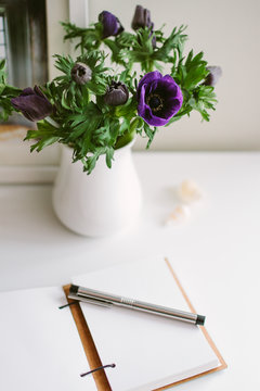 A jug of anemones on a white table with a journal open at a blank page.