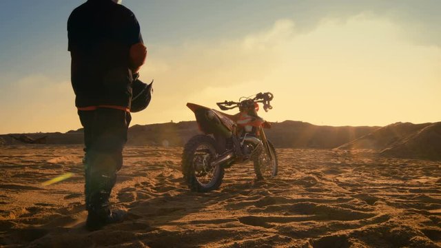 Low Angle Shot of the Professional Motocross Driver Putting on His Helmet and Getting Ready to Ride His FMX Dirt Bike on the Quarry Sand Terrain. Shot on RED EPIC-W 8K Helium Cinema Camera.