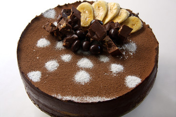 birthday chocolate and banana mousse cake decorated with cocoa powder, bananas, chocolate bars and candies on light background