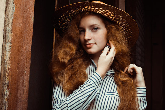 Beautiful redhead woman with long curly hair in straw hat posing near the old door