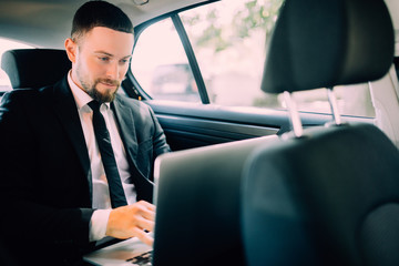 Handsome businessman sitting with laptop on the backseat of the car