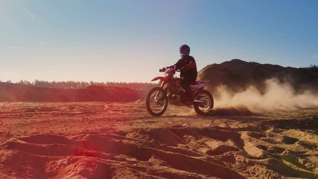 Side View Footage of the Professional Motocross Motorcycle Rider Driving on the Dune and Further Down the Off-Road Track. It's Sunset and Track is Covered with Smoke/ Dust/ Dirt. 4K UHD.