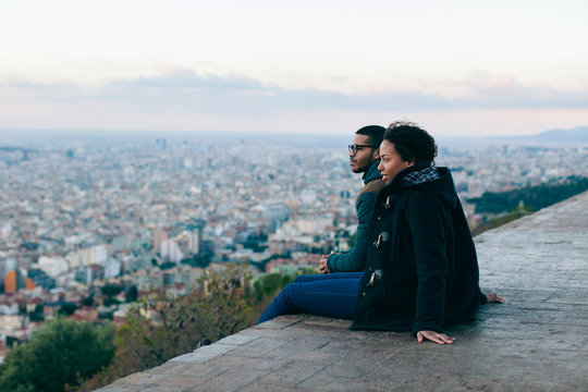 Side View Of Couple Sitting Above City At Sunrise.