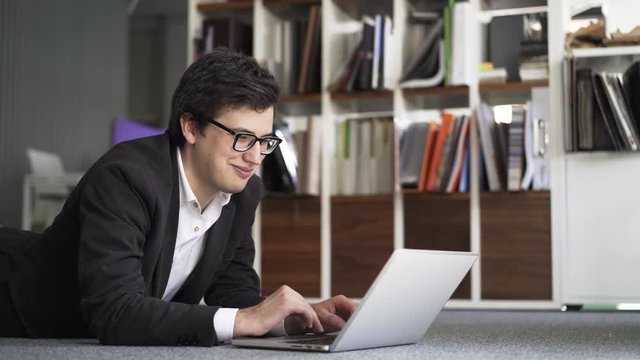 Side view of a young businessman wearing a suit and glasses lying on an office floor near a bookcase and working on his laptop. Handheld real time close up shot