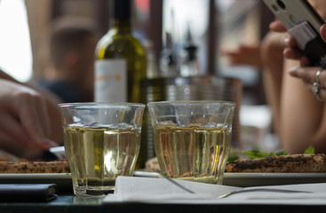 Two pizzas and glasses of white wine are on а table in a cafe in front of people. Female hands hold а phone. Concept: popular meeting place is a cafe and restaurant. 