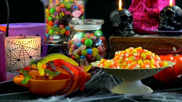 Happy Halloween trick or treat party table with bowls and apothecary jars of candy with skull candles against a black background, panning across and up.