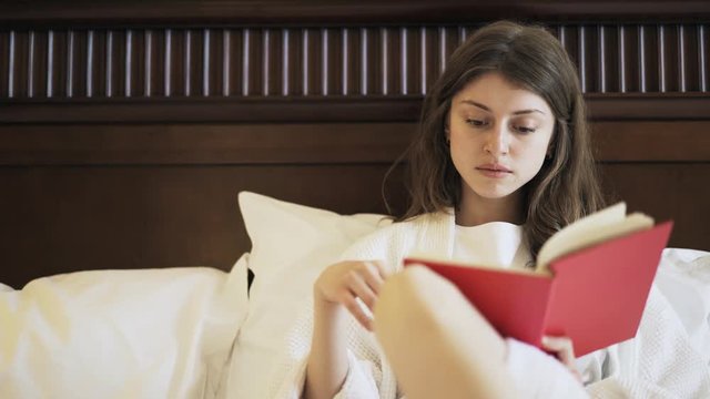 Front view of a young woman in a white bathrobe is sitting in bed and reading a book. Handheld real time medium shot