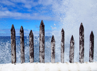 powerful waves breaking over sharp wooden spikes on a white harbor wall with splashing white foam with blue sky and clouds with the sea in the background