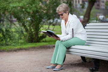 An elderly woman enthusiastically reading a book in the park on a bench, copy space