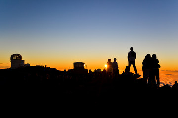 Fototapeta na wymiar Tourists watching the spectacular sunset on the summit of Haleakala Crater (10,023 ft) on the island of Maui, Hawaii. Sun setting next to a spectator. Observatories in the background. 