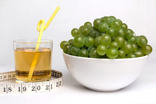 Full bowl of green grapes, a glass of grape juice with a straw and measuring tape on a white background.