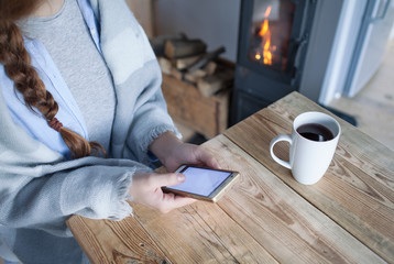 Woman with cup of coffee sitting by the fireplace and wooden table texting on the phone