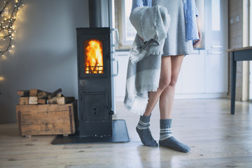 Young woman wearing winter socks standing home holding a blanket by the fireplace. Wooden cabin interior. 
