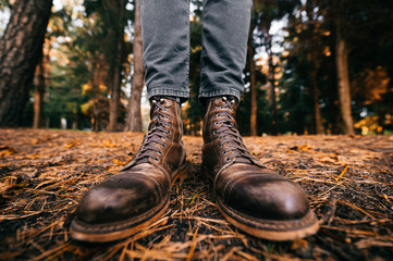 Man`s legs in woodcutter vintage leather boots and cropped jeans standing in autumn forest on ground. Fall colors and mood concept. Comfortable shoes. Walking on weekend on nature. Lens distortion.