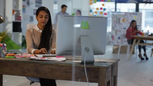 Designer working at her desk in creative office with colleagues working in background.