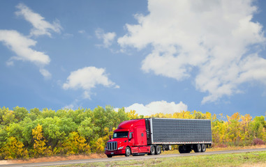 horizontal image of a semi truck with a red cab driving down the highway with beautiful fall coloured trees under a nice blue sky with white clouds on a warm fall sunny day with copy space.