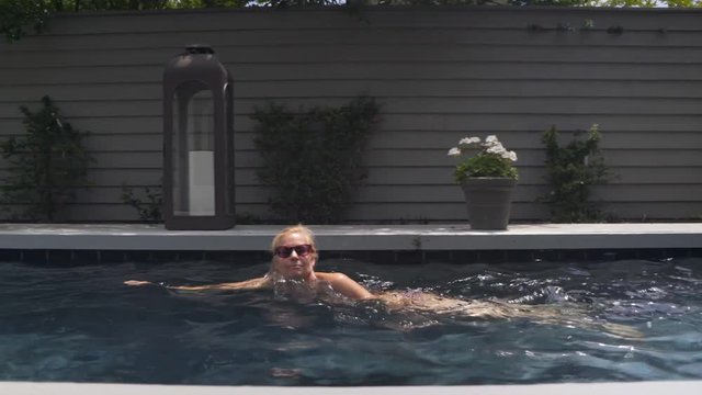 Beautiful, blonde, slim, caucasian woman wearing string bikini doing side stroke in private pool lined with flowers and grey wall. Camera moves with swimmer.