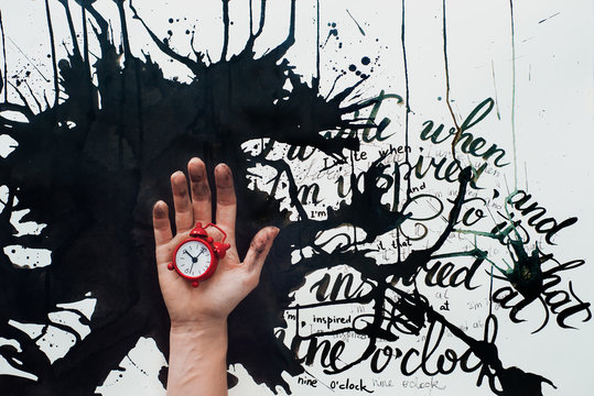 Hand holding a tiny red alarm clok on a background with spilled ink and an inspirational quote. Creative flat lay with writer workplace. Conceprual still life with copy space.