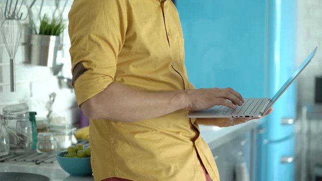 Man In Yellow Shirt Standing In His Modern Kitchen And Using Notebook
