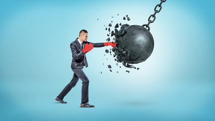 A tiny businessman in red boxing gloves breaks a large swinging wrecking ball in small pieces.