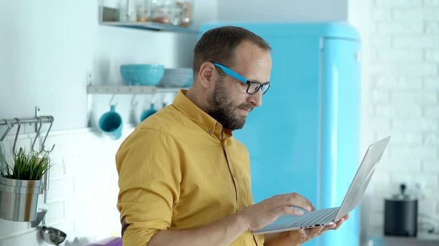 Handsome Man In Yellow Shirt Standing In His Modern Kitchen And Using Silver Notebook
