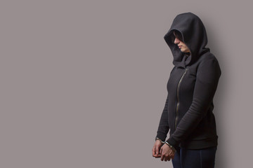 side view of a girl in a black hood with handcuffed hands isolated on a gray - obrazy, fototapety, plakaty