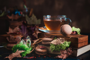Glass tea cup in autumn still life with macarons, moss, maple leaves and wooden boxes. Dark food photography with sweets.