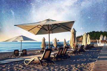 Scenic view of sandy beach on the beach with sun beds and umbrellas open against the sea and night sky. Fantastic starry sky and the milky way
