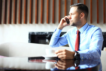 Handsome man is working in cafe, drinking cup of coffee, smiling and looking at smart phone.