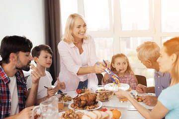 The family eats at the festive table for Thanksgiving. A woman is laying out food, her family is already eating