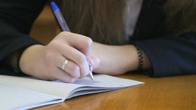 Schoolgirl dressed in a black suit sits at a school desk writing text in exercise book using ballpoint pen. Close-up