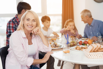 A woman is posing at the table, after which her family is eating. They celebrate Thanksgiving Day