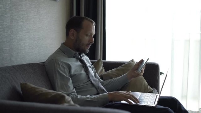 Young businessman working with tablet and laptop sitting on sofa at home
