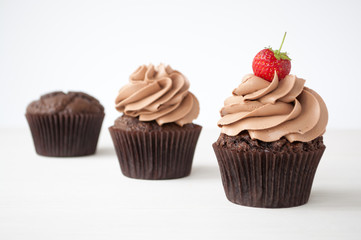 Cupcakes with whipped chocolate cream, decorated fresh strawberry on white wooden table. Picture for a menu or a confectionery catalog, showing different stages of creation.