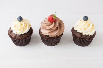 Cupcakes with whipped chocolate and vanila cream, decorated fresh strawberry, blueberry, gold confectionery sprinkling on white wooden table. Picture for a menu or a confectionery catalog.