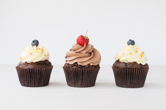Cupcakes with whipped chocolate and vanila cream, decorated fresh strawberry, blueberry, gold confectionery sprinkling on white wooden table. Picture for a menu or a confectionery catalog.