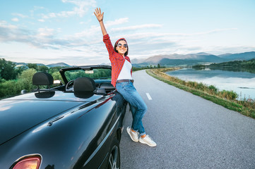 Happy smiling young woman stay near the cabriolet car on the mountain road