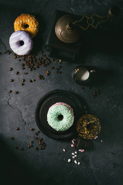 Variety of colorful glazed donuts with chopped chocolate, pink sugar, coffee beans vintage coffee grinder on black texture background. Dark rustic still life. Flat lay with space