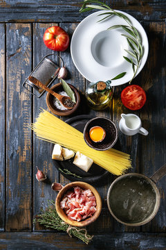 Ingredients for traditional italian pasta. Uncooked spaghetti, tomatoes, pancetta bacon, parmesan cheese, egg yolk, salt, pepper, empty plate, pan water, over old plank background. Top view with space