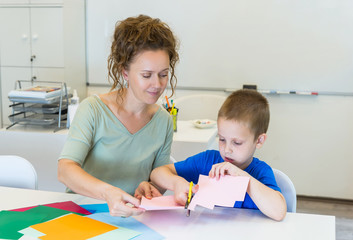 teacher woman and child boy cutting collage color paper with scissors in the classroom