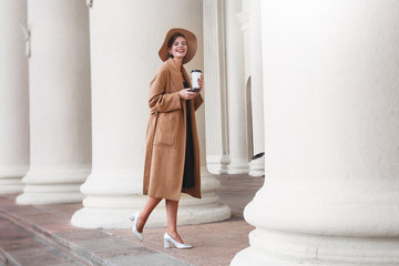 Girl in a brown coat a brown hat is walking and posing in the city interiors. The girl is smiling, checking her smartphone and drinking coffee from take away cafe. The photo looks very positive cause