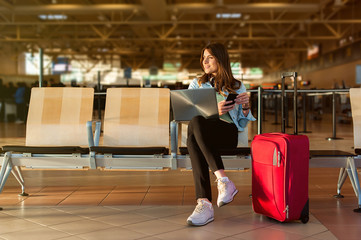 Airport Young female passenger on smart phone and laptop sitting in terminal hall while waiting for...