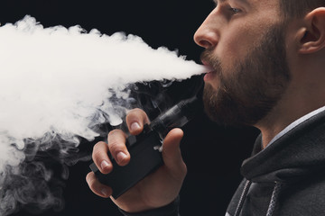 Young man vaping e-cigarette with smoke on black