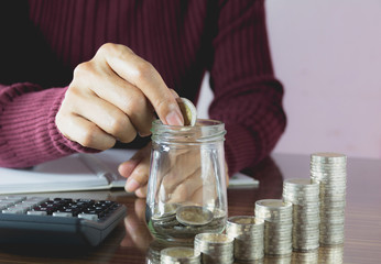 Woman process and drop coin into the glass with stack coins and calculator. Woman working on table.