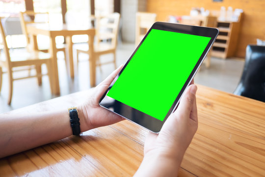 Mockup image of hands holding black tablet pc with white blank green screen on wooden table in modern cafe