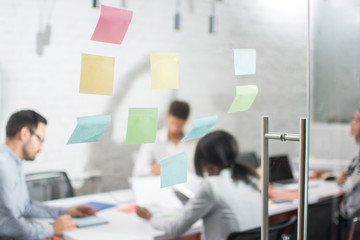 Selective focus image of blank adhesive notes on transparent glass wall in office. Blurred business people having meeting in the background.